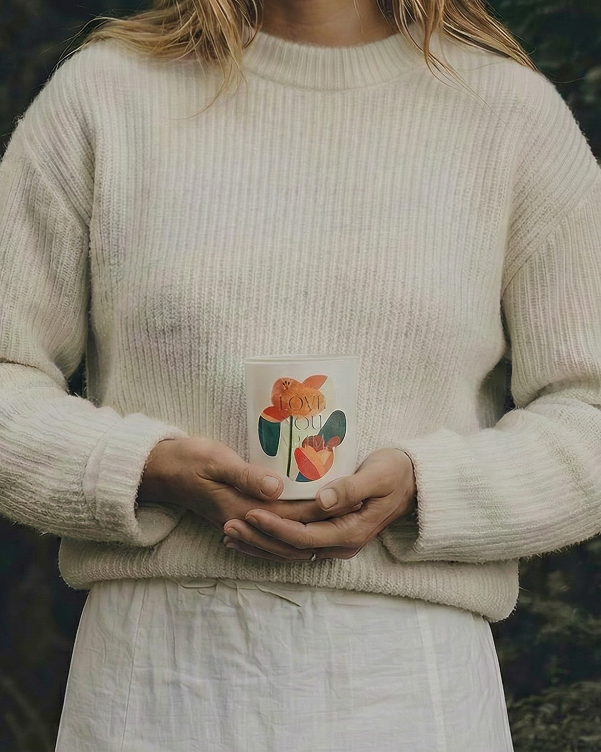 Person wearing a beige sweater holding a mug with a floral design against a blurred natural background