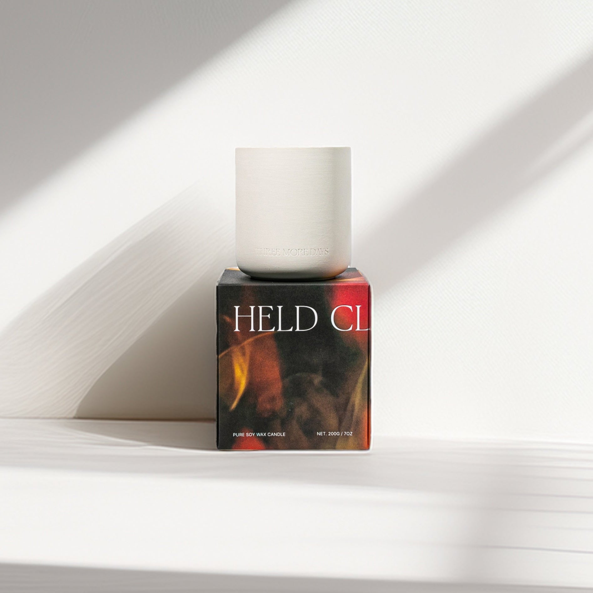 Candle in a box labeled 'HELD CL' on a white surface with light shadows