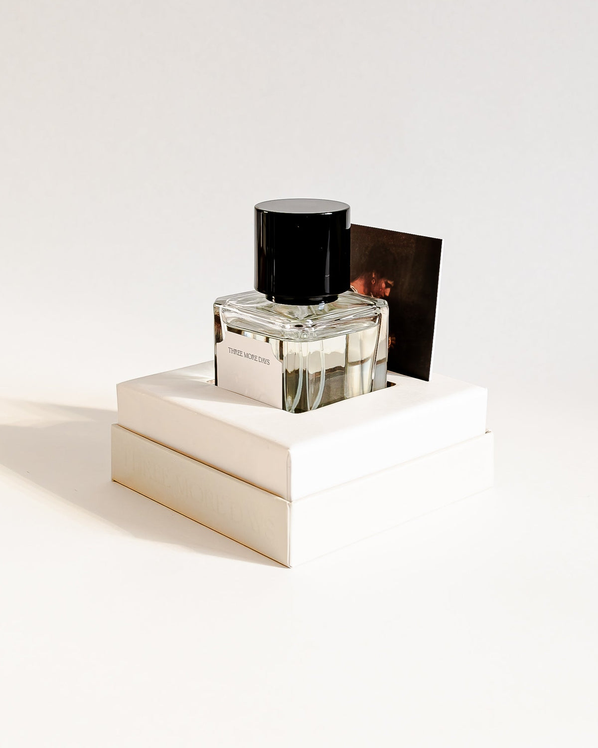 A clear glass perfume bottle with a black cap, displayed on top of two stacked cream-colored boxes against a clean white background.