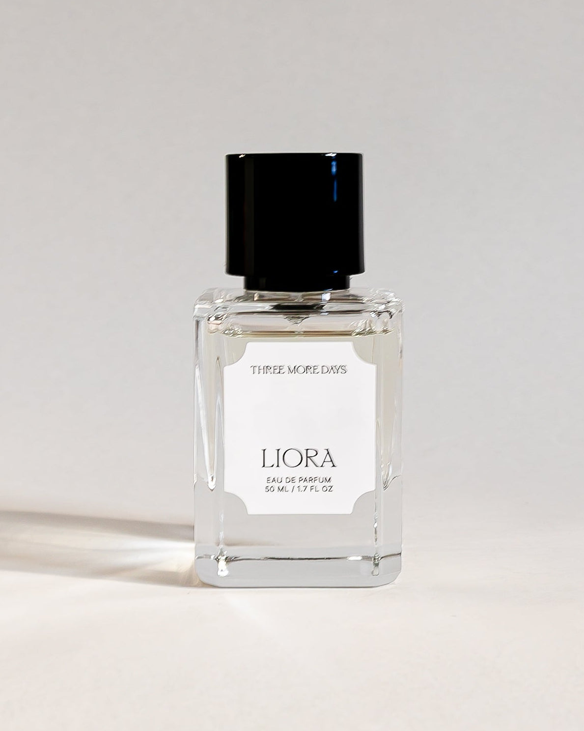 A small clear glass perfume bottle with a black cap and a white label reading 'L'ORLA' and 'EDCOTTE', standing on a white studio surface with a curved white backdrop behind it.