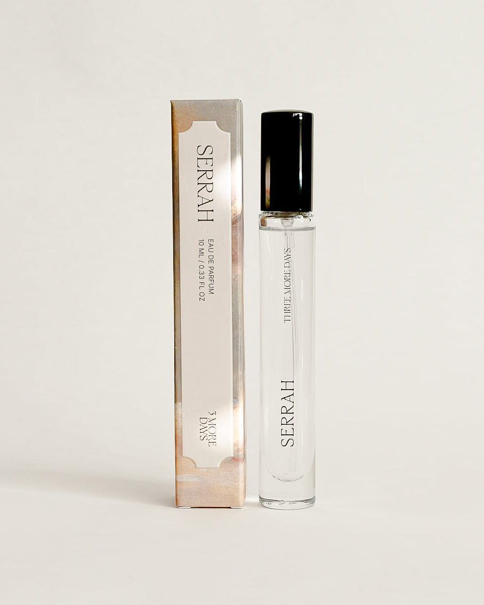 A small clear glass perfume roller bottle with a black cap stands next to its tall rectangular box on a white draped studio backdrop. The packaging has a warm beige/champagne tone with black text, and the bottle itself is transparent with a black cap.