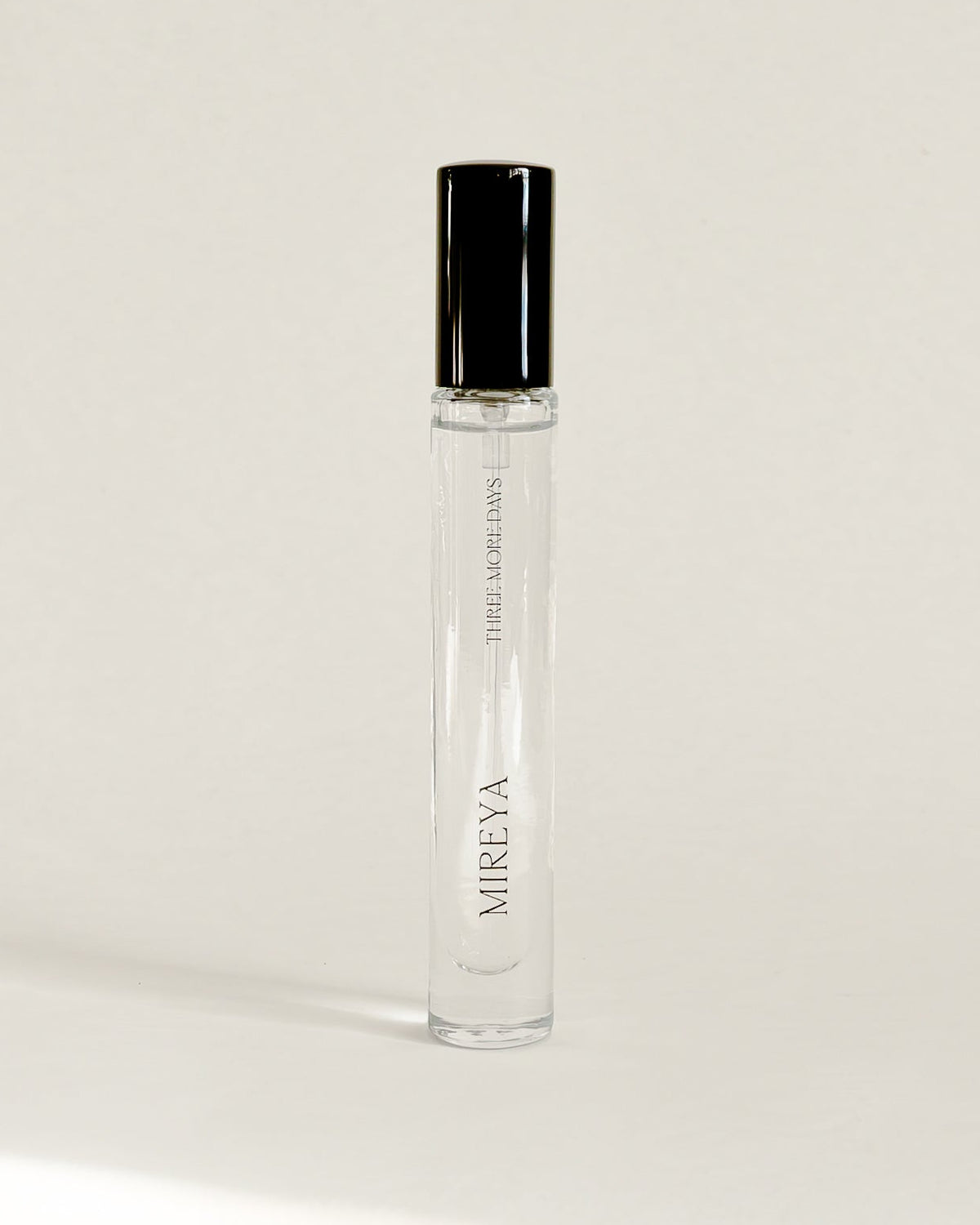 A clear glass perfume roller bottle with a black cap, standing upright on a white surface against a white backdrop.