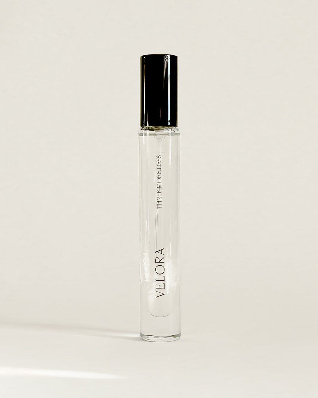 A clear glass perfume roller bottle with a black cap, standing upright on a white surface against a white backdrop.