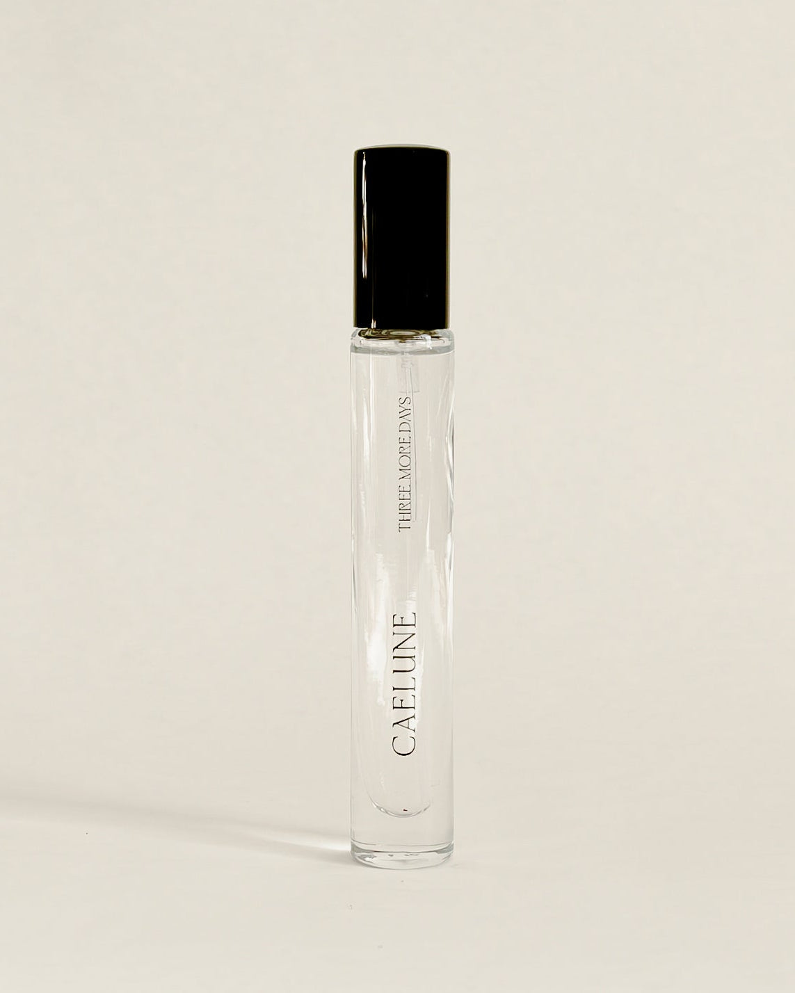 A clear glass perfume roller bottle with a black cap, standing upright on a white surface against a white backdrop.