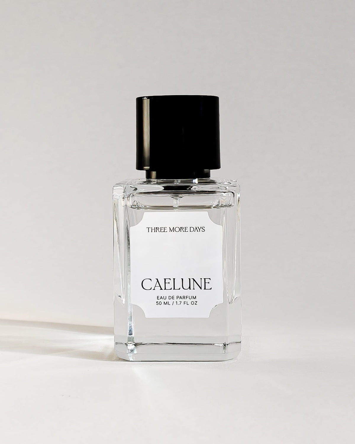 A clear glass perfume bottle with a black cap and a white label reading 'CAELLINE' and 'SUPER ALCHEMY', set against a white studio backdrop.