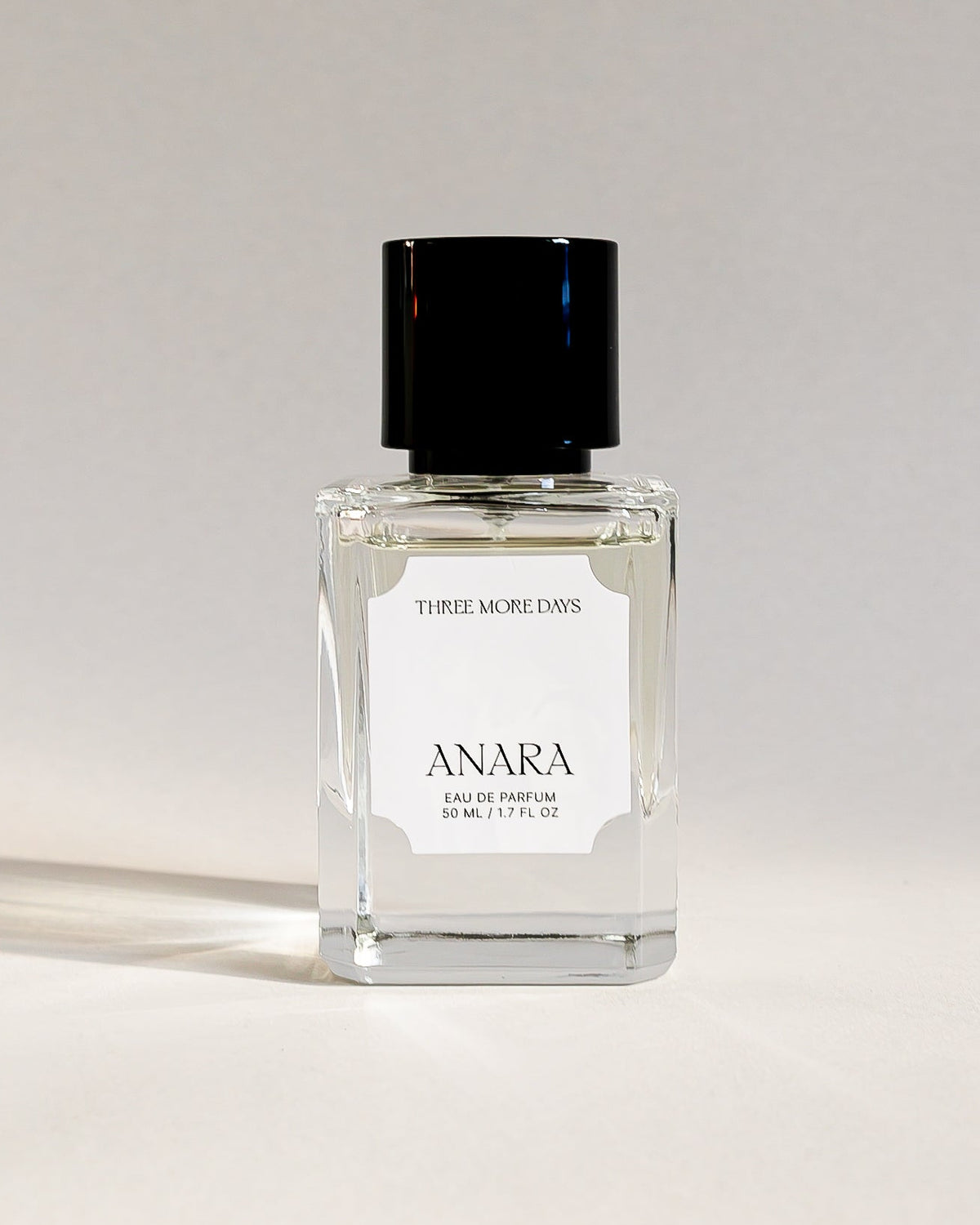 A clear glass perfume bottle with a black cap and a white label reading 'ANARA' and 'FUMIGRIS ANORA', photographed on a white studio background with a visible shadow.