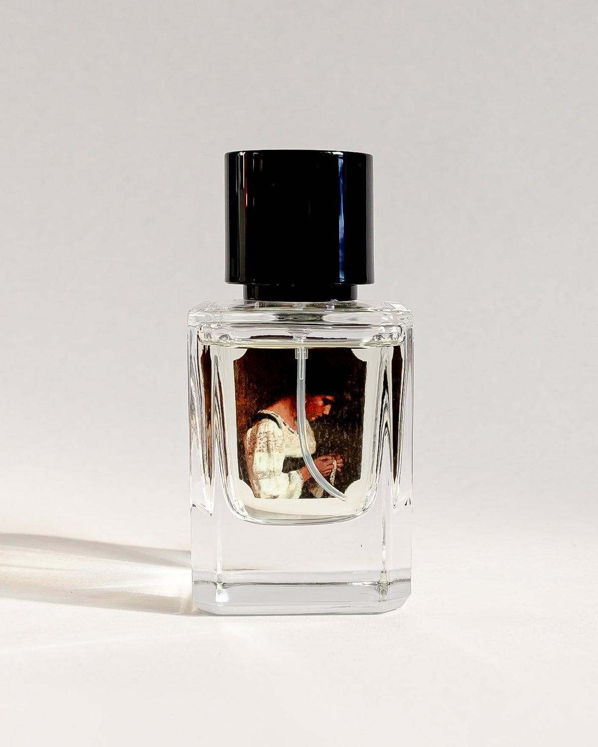 A small clear glass perfume bottle with a black cap, standing on a white surface against a white background.