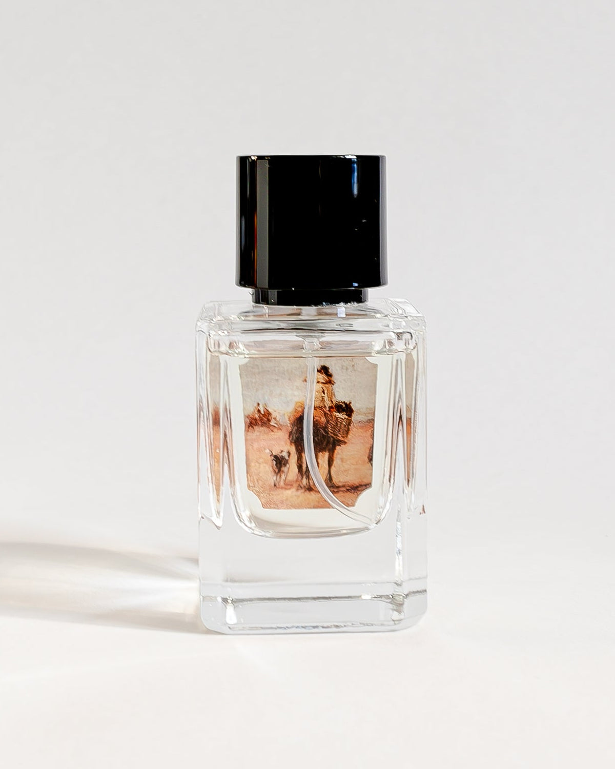 A small clear glass perfume bottle with a black cap and a printed label, standing on a white surface against a white curved backdrop.