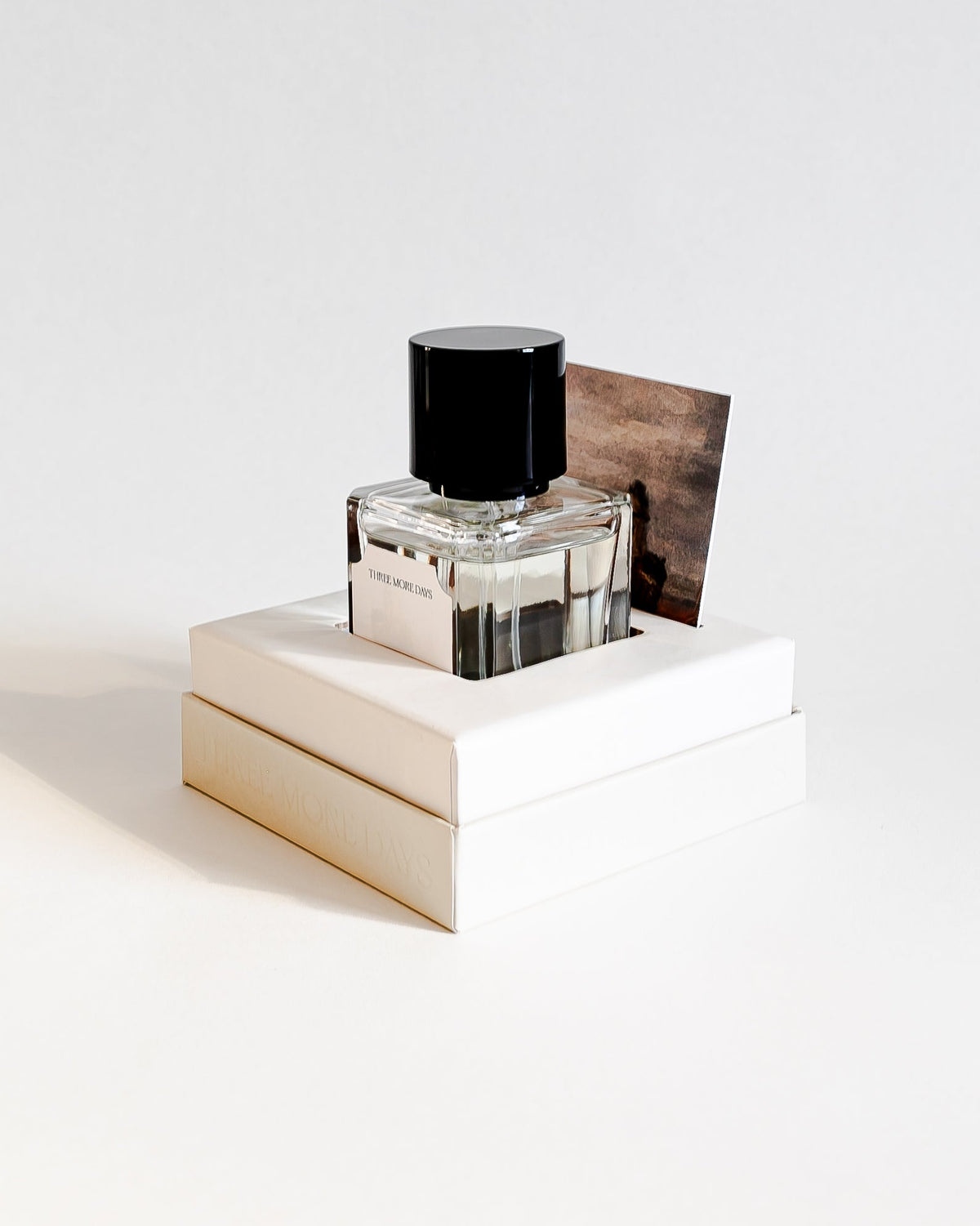 A clear glass perfume bottle with a black cap, displayed on top of two stacked white boxes against a clean white background.
