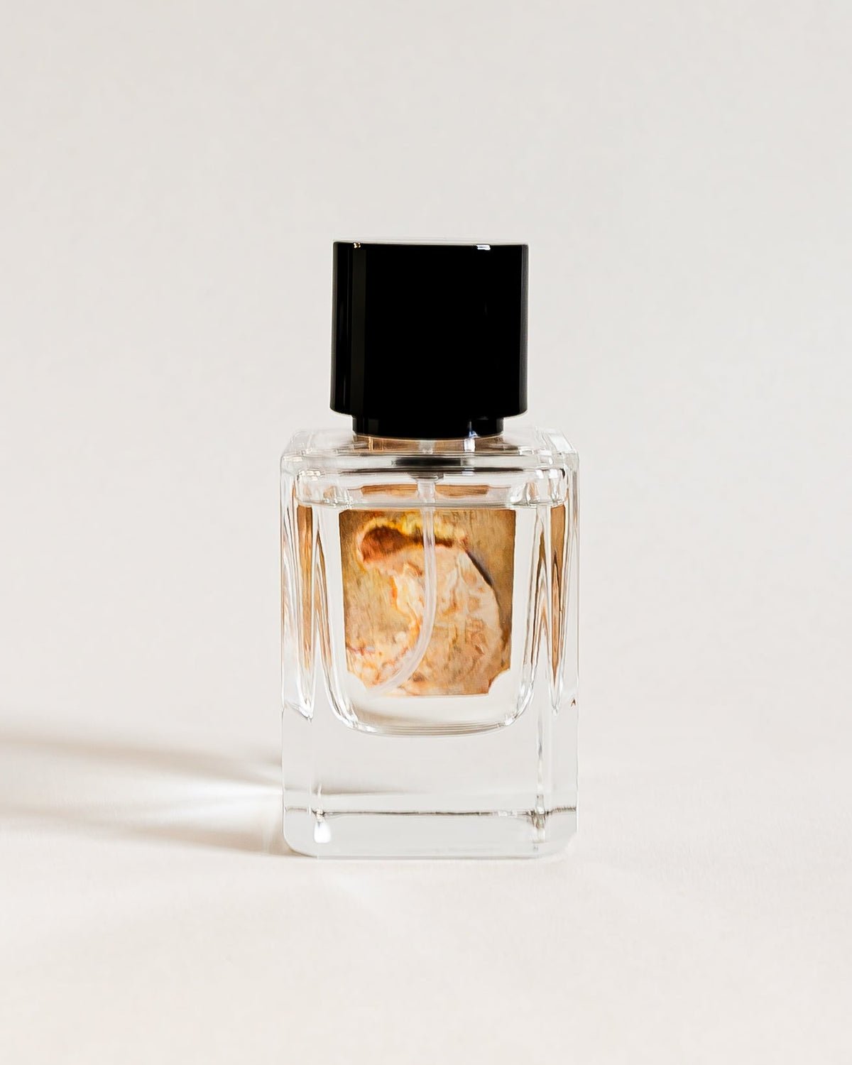 A small clear glass perfume bottle with a black cap and a gold-toned label, centered on a white studio backdrop.