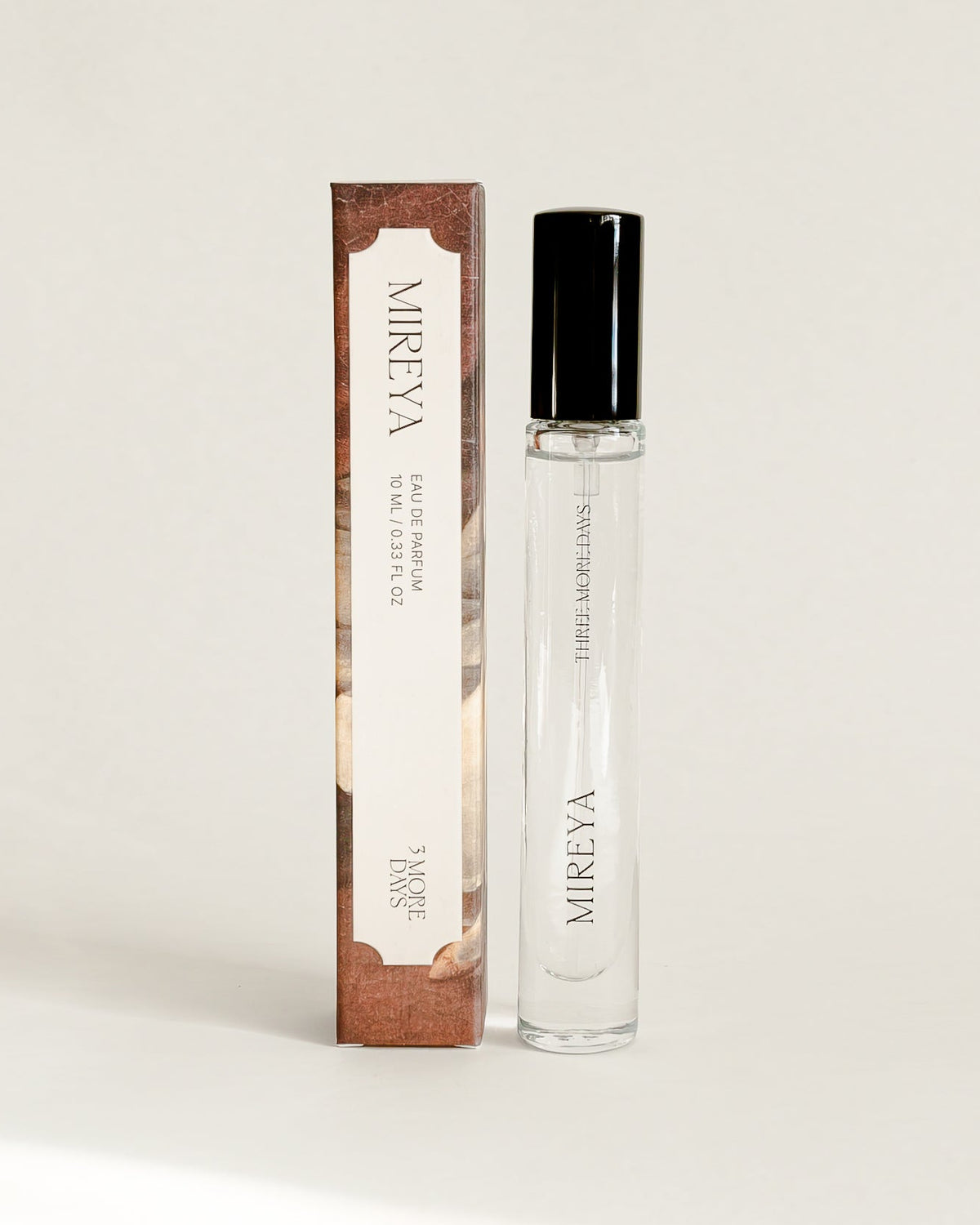 A small cylindrical cosmetic bottle with a black cap stands next to its matching rectangular box on a draped white paper backdrop.