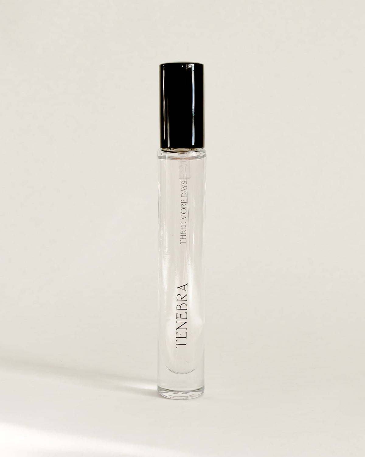 A clear glass perfume roller bottle with a black cap, standing upright on a white surface against a white backdrop. 