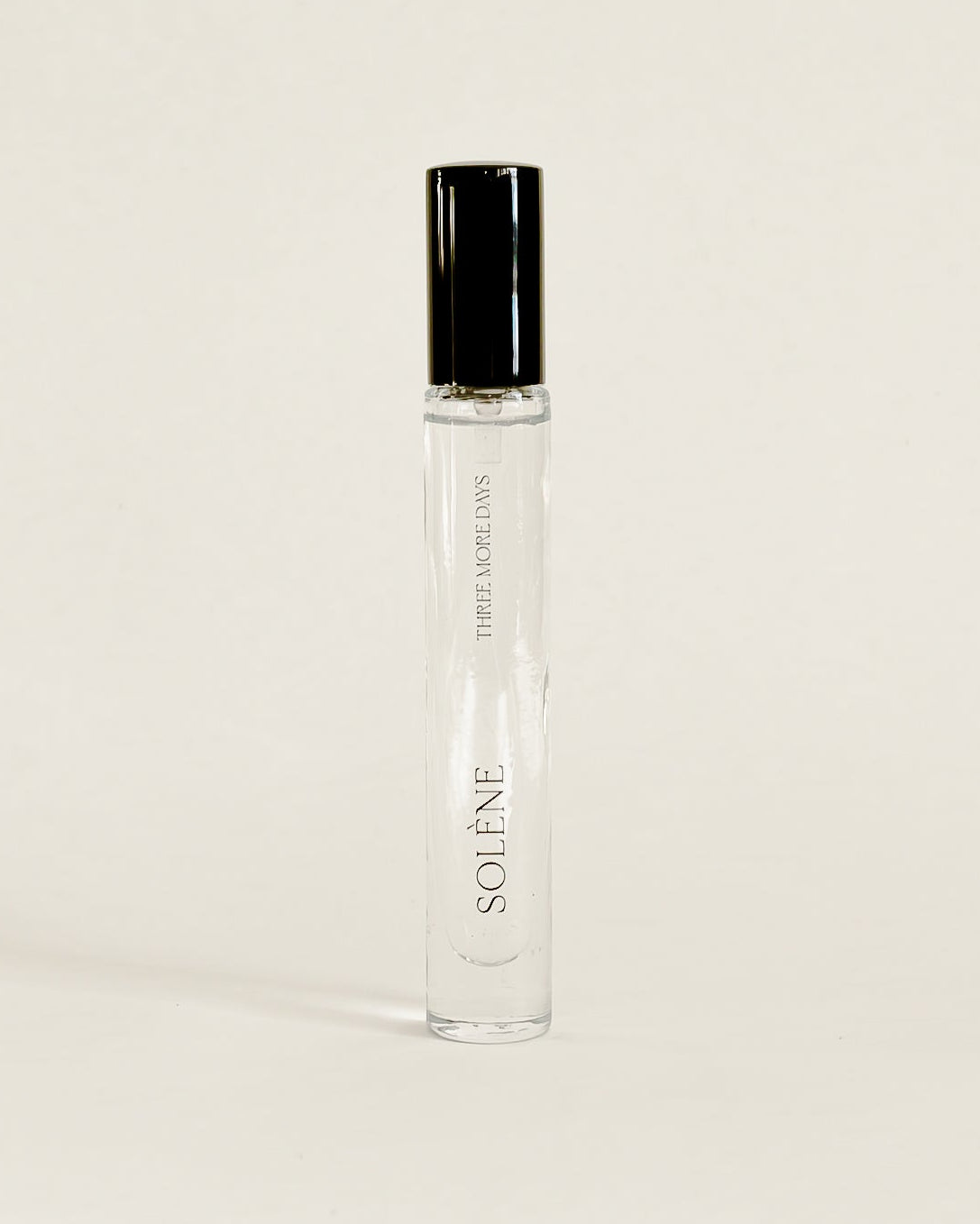 A clear glass perfume roller bottle with a black cap, standing upright on a white surface against a white backdrop. 