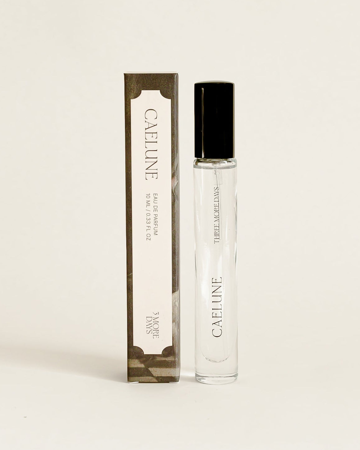 A clear glass perfume roller bottle with a black cap stands next to its tall rectangular box packaging, both featuring the brand name.